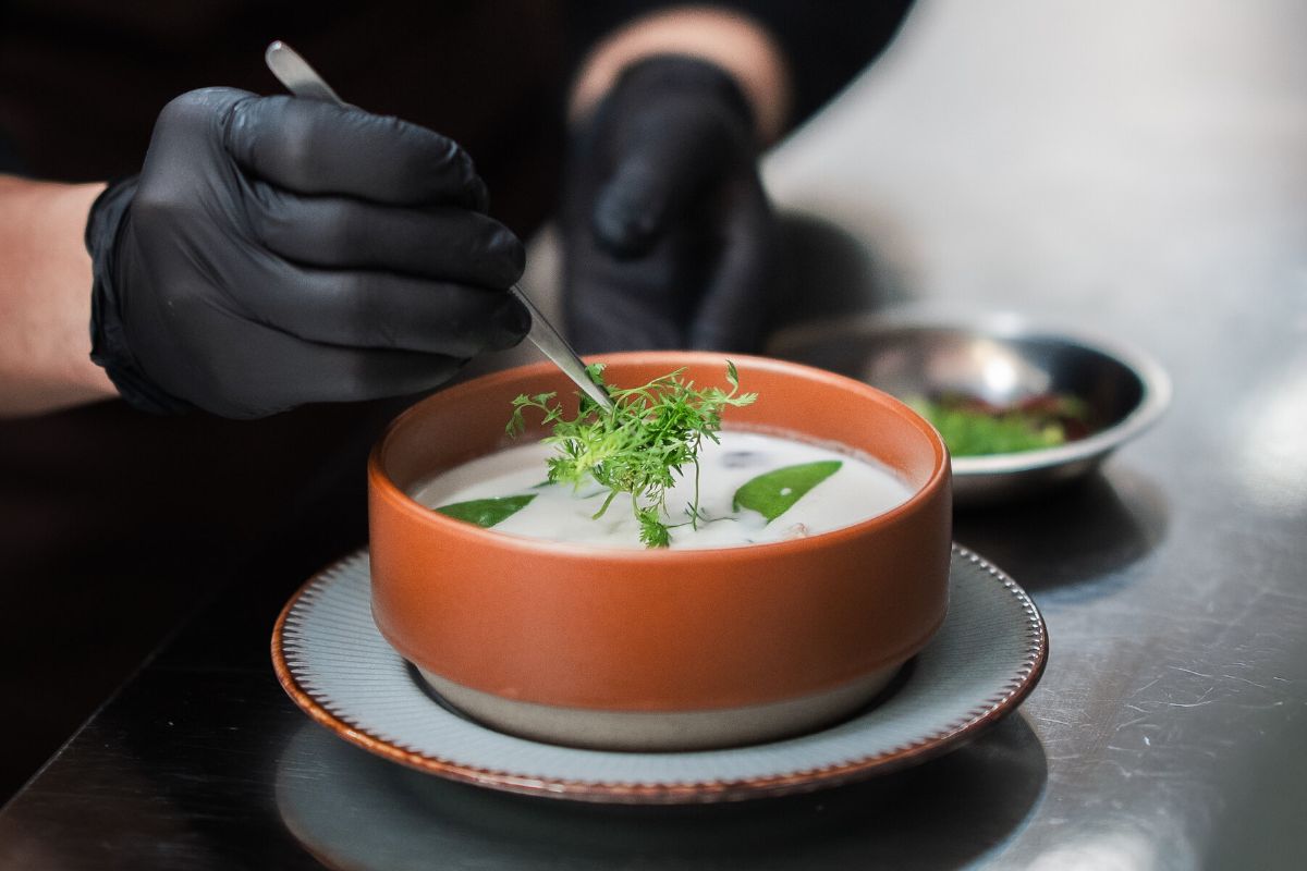 A chef in black uniform garnishing a bowl of pale soup with fresh green herbs in the WIROON kitchen