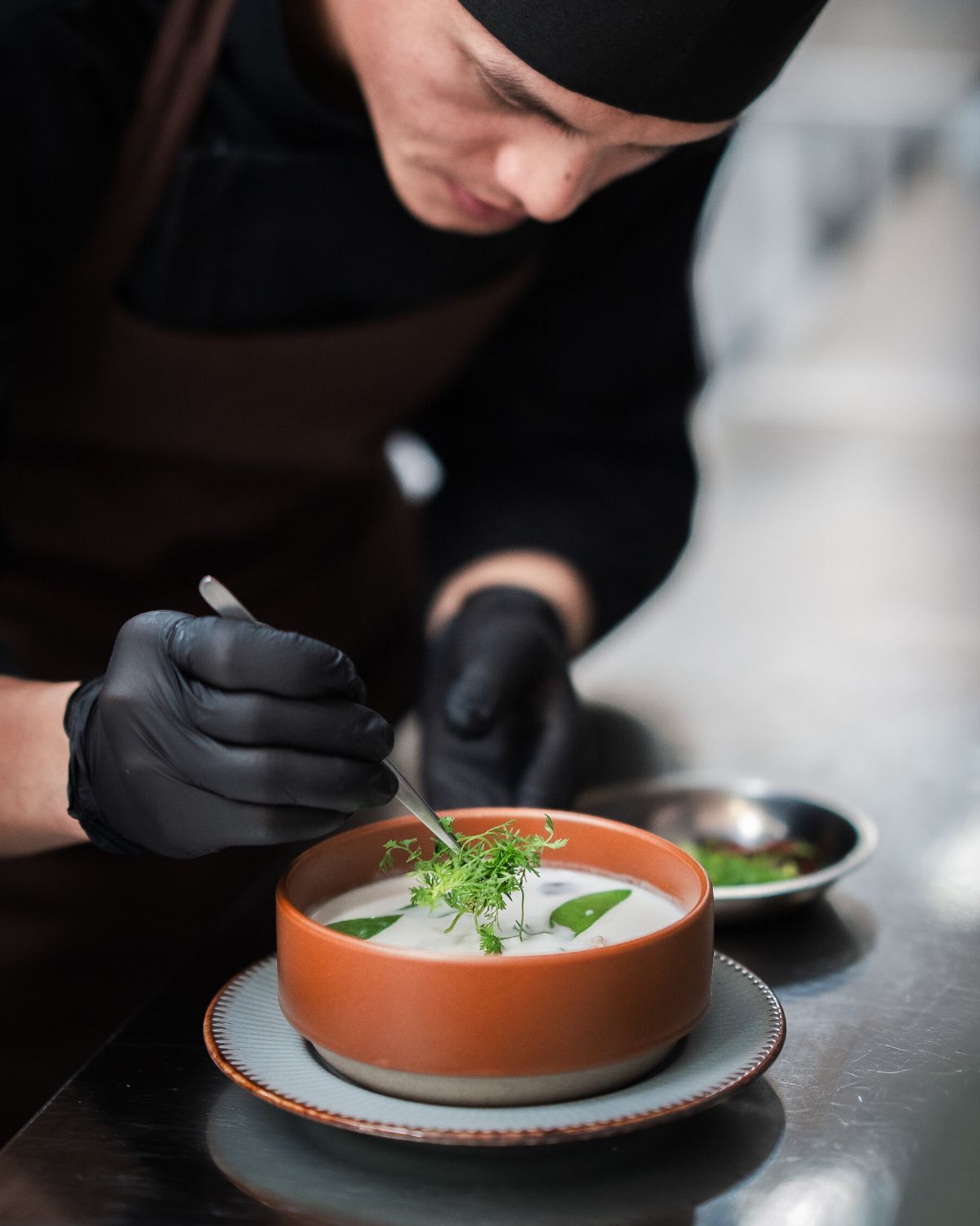 A chef in black uniform garnishing a bowl of pale soup with fresh green herbs in the WIROON kitchen