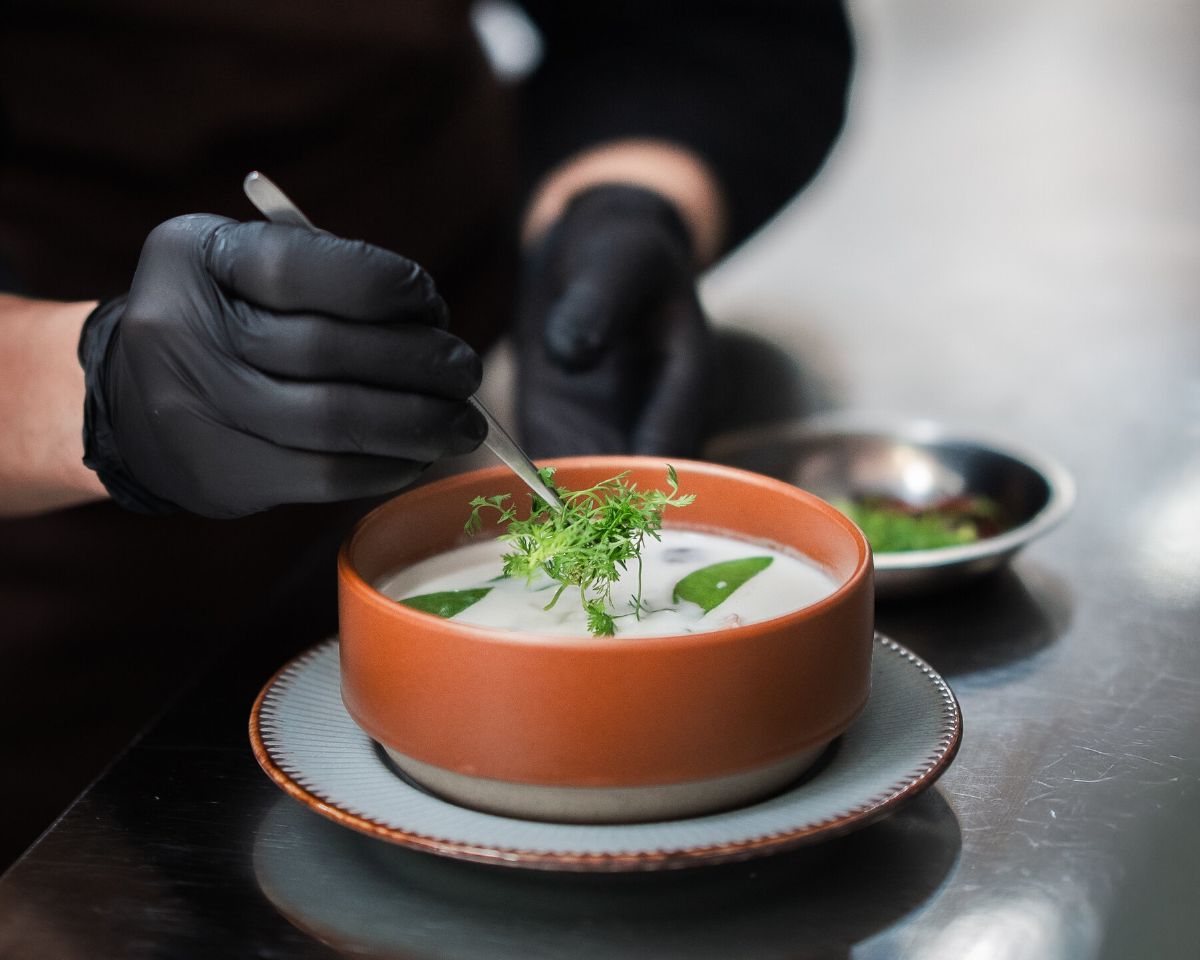 A chef in black uniform garnishing a bowl of pale soup with fresh green herbs in the WIROON kitchen