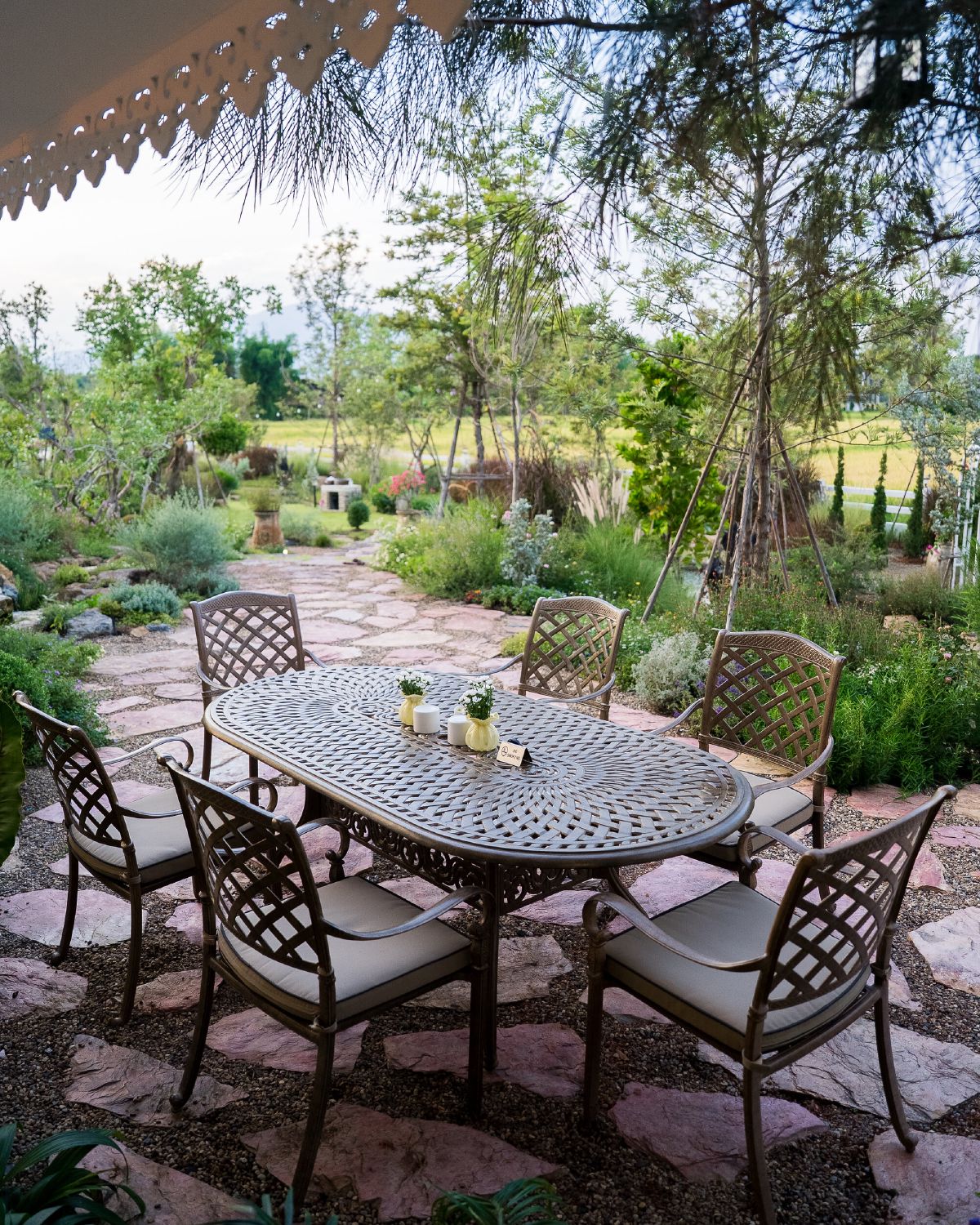 An oval metal garden table laid with small vases, surrounded by six cushioned chairs on a stone terrace, with planted borders and open fields beyond