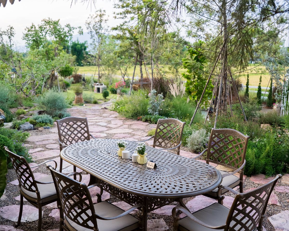 An oval metal garden table laid with small vases, surrounded by six cushioned chairs on a stone terrace, with planted borders and open fields beyond