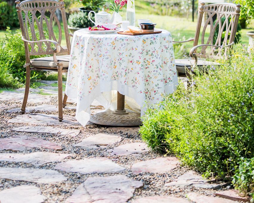 A small round table set with a floral cloth for breakfast — teapot, bread and fruit — among garden greenery in morning light