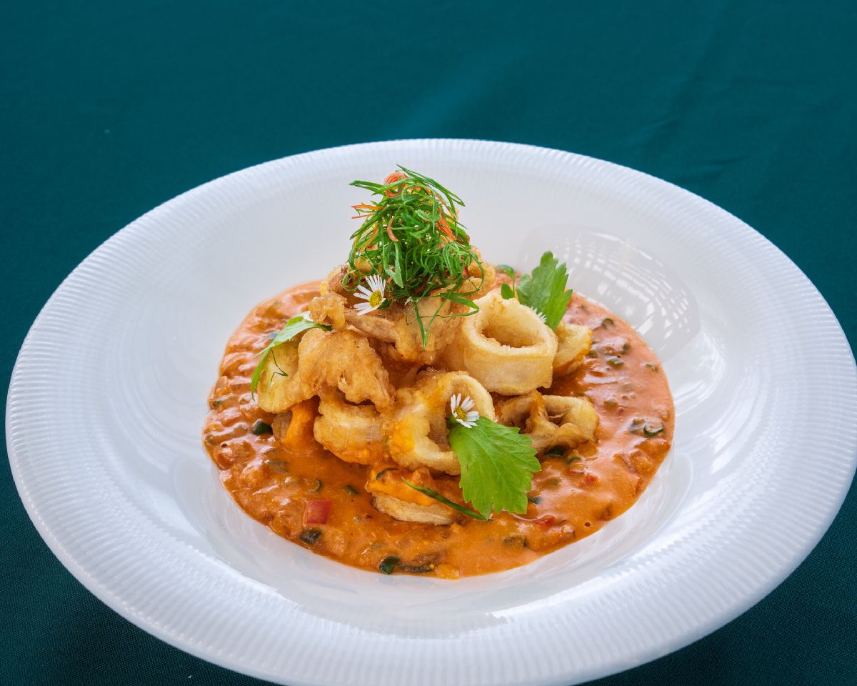 A plated dish of crisp-fried seafood in a red curry sauce, topped with herbs and edible flowers, on a white plate against teal linen