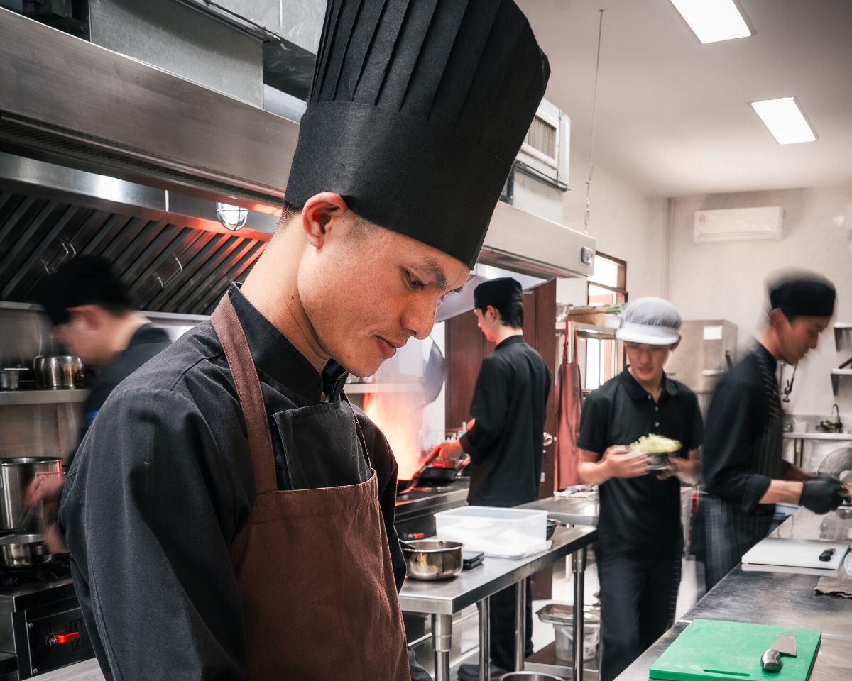A chef in the WIROON kitchen plating a dish into an earthenware bowl, surrounded by bowls of fresh tomatoes, lemongrass and citrus