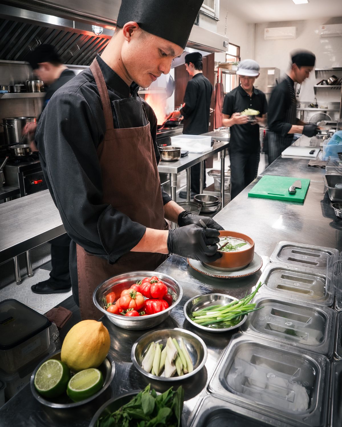 A chef in the WIROON kitchen plating a dish into an earthenware bowl, surrounded by bowls of fresh tomatoes, lemongrass and citrus